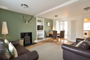 A living room with a fireplace, leather sofas, a dining area, and a kitchen visible through an open door.