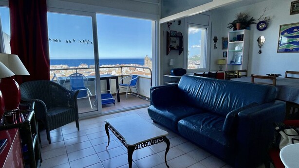 Ein Wohnzimmer mit blauem Sofa, einem kleinen Tisch und einem Sessel. Ein Balkon bietet Blick auf das Meer und Segelboote.