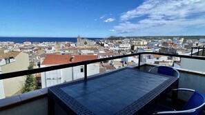 Un balcon avec une table et des chaises, donnant sur une ville côtière avec ses bâtiments et son église.