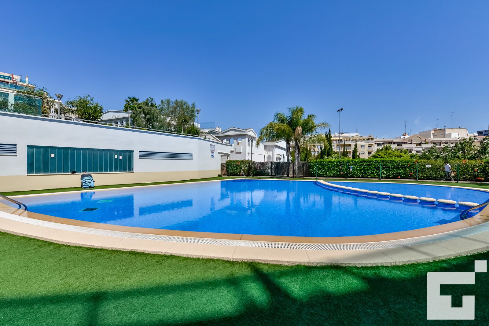 A swimming pool with a clear blue water surface, surrounded by a green lawn and a white building with large windows.