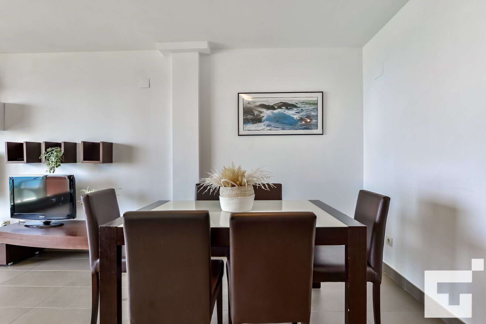 A modern dining area with a white table, six brown chairs, a decorative plant, and a framed picture on the wall.