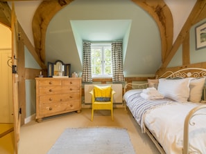 A bedroom with a wooden dresser, a yellow chair, a bed with white bedding, and a window with checkered curtains.