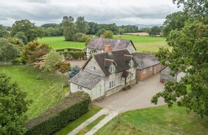 Een groot, traditioneel huis met meerdere schoorstenen, omgeven door uitgestrekte groene velden en bomen.