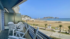 Un balcon avec des chaises et une table en plastique blanc, d’où l’on a une vue dégagée sur une plage, la mer et les bâtiments situés au loin.