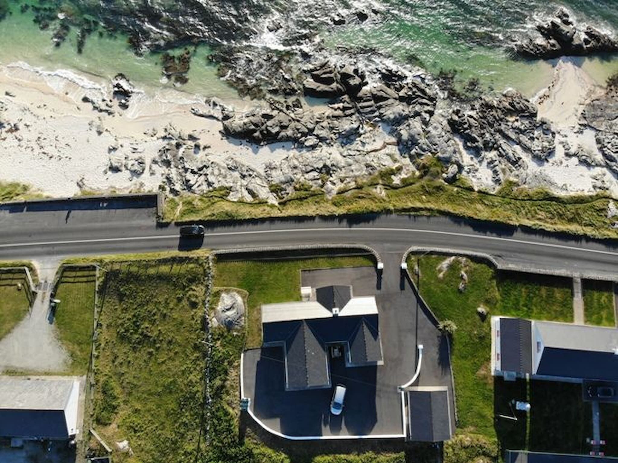 An aerial view of a coastal property with a road, a parking area, and a house with a garage.