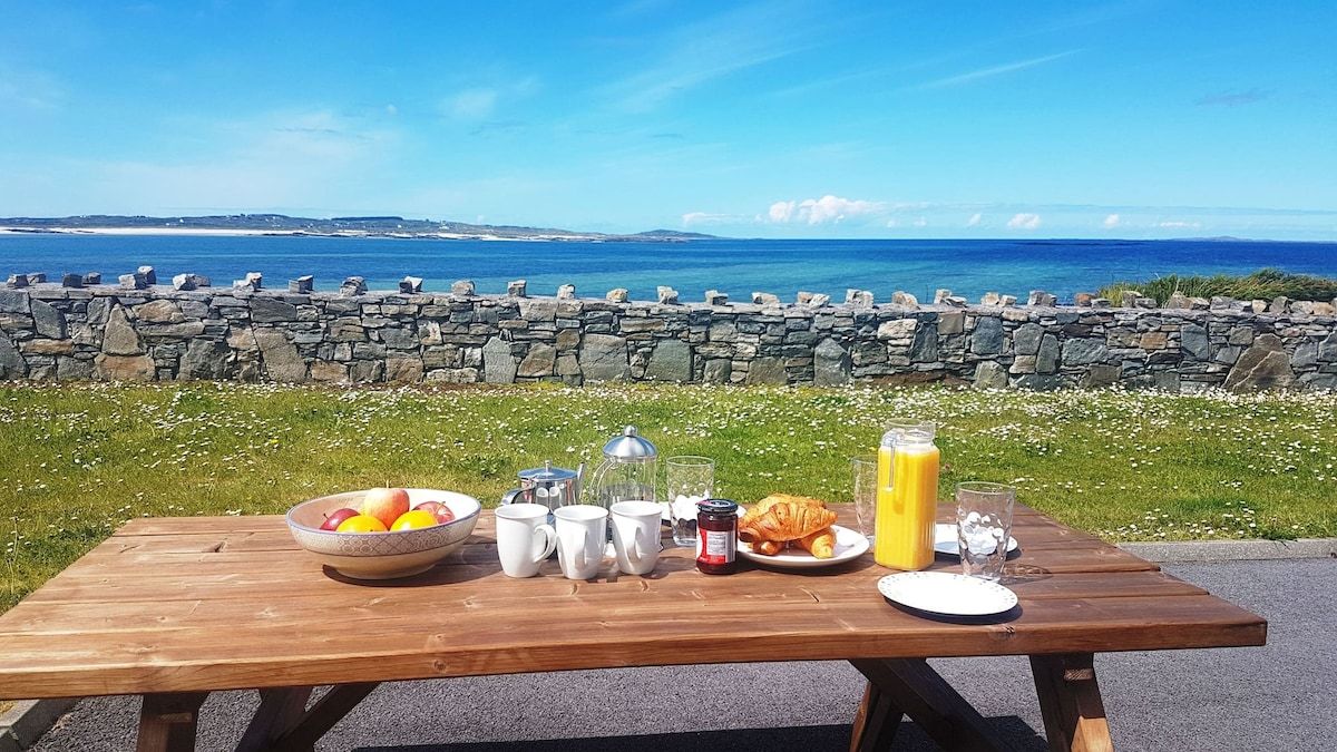 A picnic table with breakfast items, a stone wall, and a view of the sea.