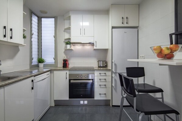 Een compacte keuken met witte kastjes, een inbouwoven, een koelkast en een kleine eethoek met twee stoelen.