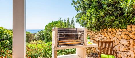 An outdoor grill area with a stone wall and a bench, overlooking a garden and the sea.