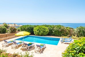 Piscina con sillas de descanso y una sombrilla amarilla, rodeada de vegetación y con vistas al mar.
