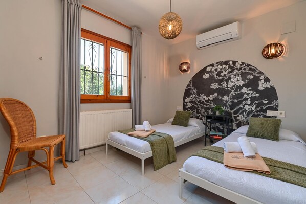Una habitación de hotel con dos camas individuales, una silla de mimbre, un ventanal con cortinas y un aire acondicionado instalado en la pared.
