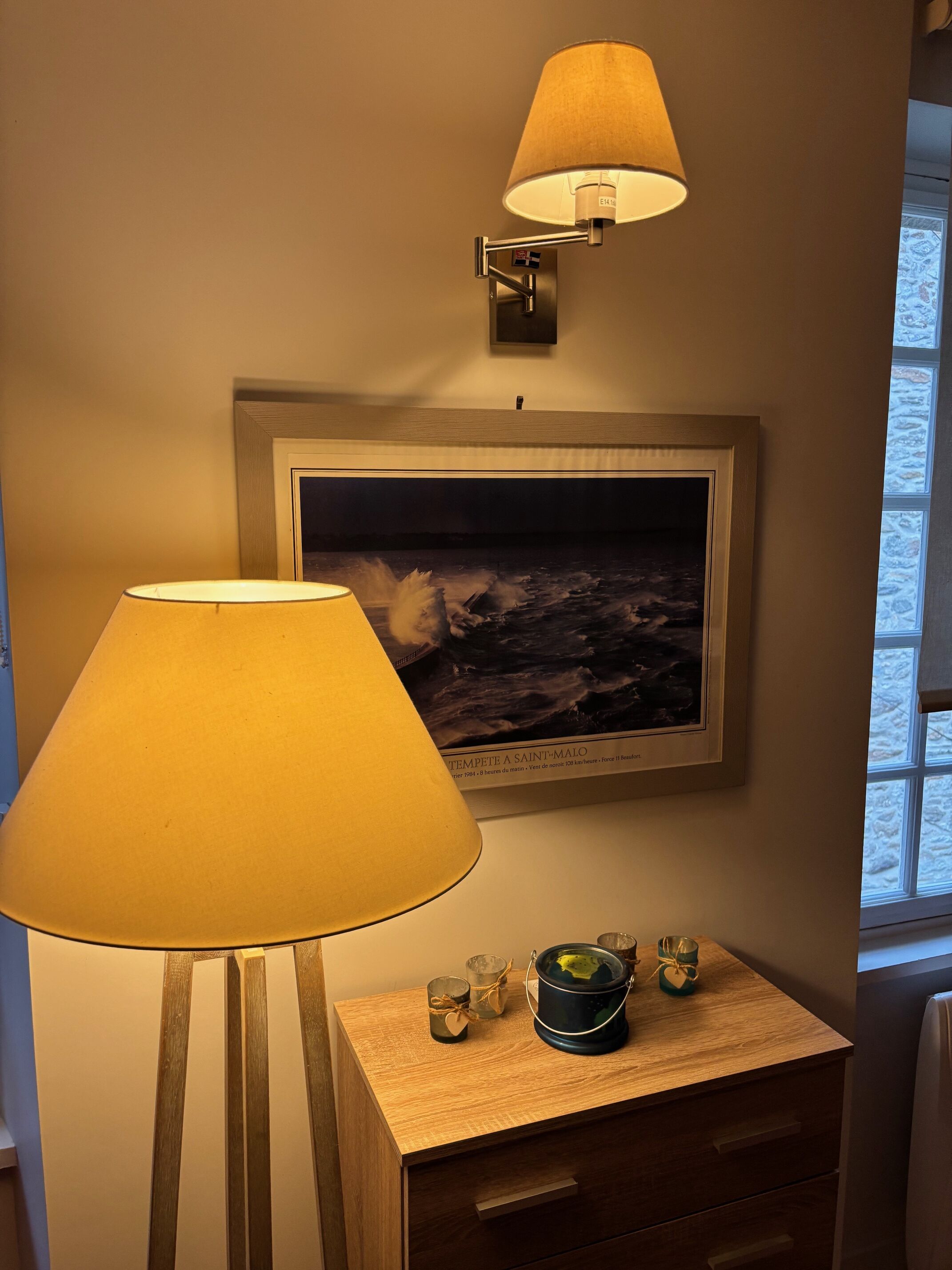 A room with a wooden nightstand, a lamp, a framed picture of a stormy sea, and a window with a view of snow.