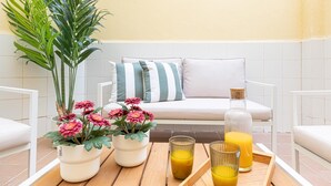 Une terrasse avec une table en bois, des plantes en pot, un canapé avec des coussins à rayures et une bouteille de jus d’orange.