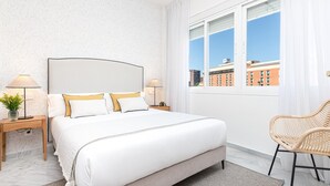 Une chambre d’hôtel moderne avec un grand lit, deux tables de chevet avec des lampes, un fauteuil en osier et une vue sur les immeubles par la fenêtre.