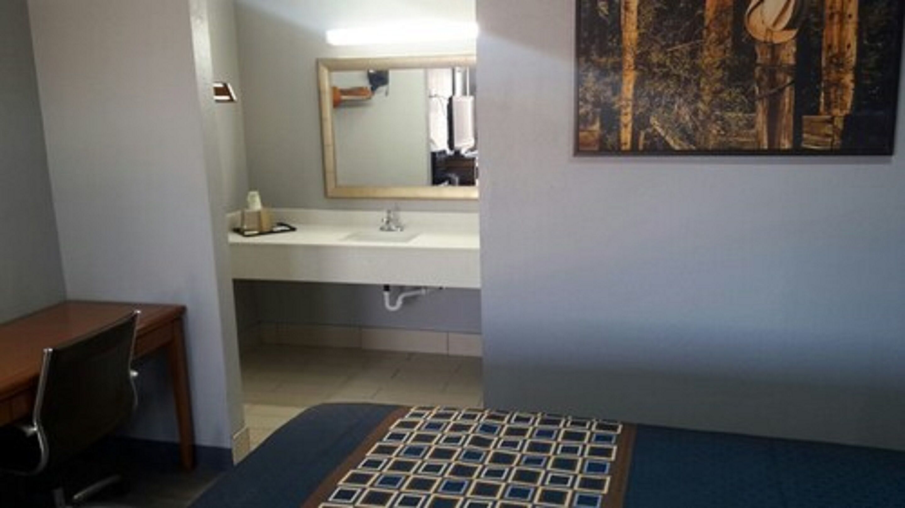 Una habitación de hotel con cama, escritorio, baño con lavamanos y espejo, y un cuadro en la pared.