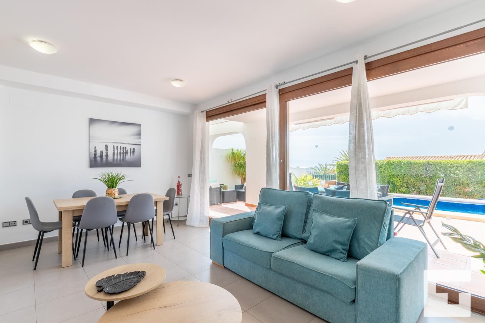 A modern living room with a sofa, dining table, and chairs. There is a large window with curtains, a view of the outdoors, and a pool area visible through the window.