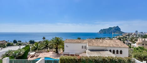 A coastal property with a swimming pool, a white building with a tiled roof, and a view of the sea and a mountain in the distance.