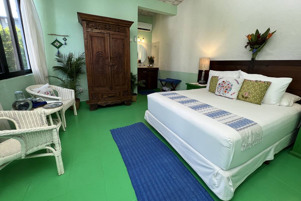 Quarto com uma cama grande, um guarda-roupa de madeira, uma cadeira de vime, uma mesinha e uma planta em vaso.