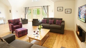 Ein geräumiges Wohnzimmer mit einer Couch, Sesseln und einem Couchtisch. Ein Kamin und ein großes Fenster mit Vorhängen sind ebenfalls vorhanden.