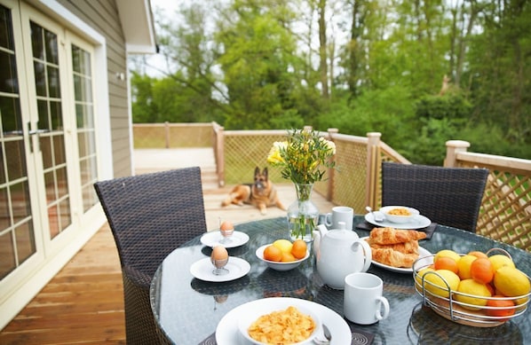 A dining table set outdoors with breakfast items, a dog, and a view of a house and greenery. - Brook Lodge holiday cottage is located just a stone’s throw away from Colchester. (Colchester)