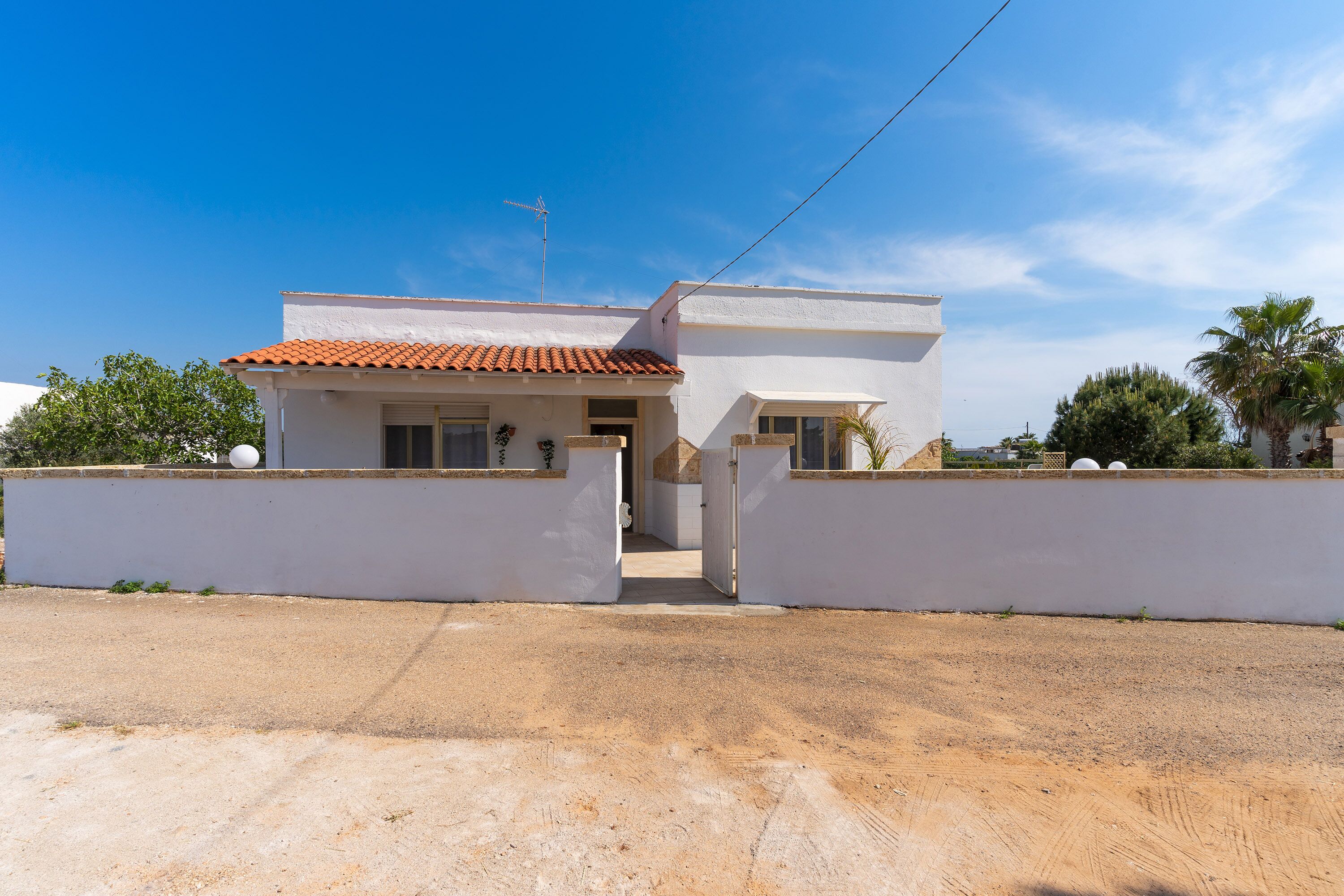 Une maison à deux étages, avec un toit recouvert de tuiles rouges, des murs blancs et une entrée fermée par un portail.