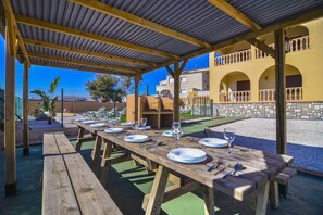 An outdoor dining area with wooden tables and benches, a covered roof, and a view of a yellow building with arches.
