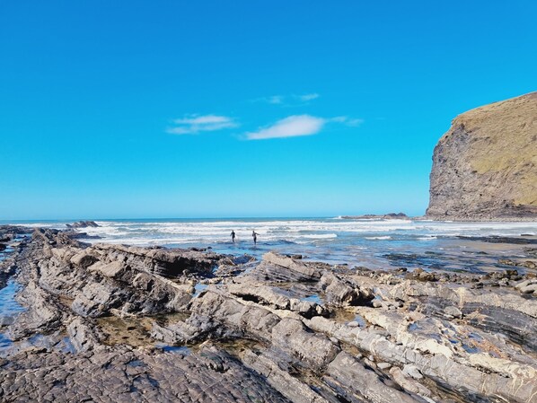 Een rotsachtig strand met twee mensen die wandelen, een heldere lucht en een klif op de achtergrond.