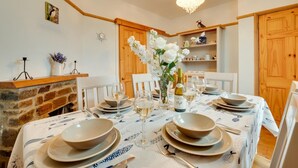 A dining area with a table set for six, featuring a white tablecloth, plates, bowls, and wine glasses. There is a vase with white flowers, a bottle of wine, and a clock on the wall.