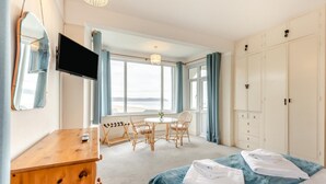A hotel room with a bed, a wooden dresser, a TV, a dining table with chairs, and a view of the beach through large windows. - 3 Bed in Woolacombe (oc-n1711) (Woolacombe)