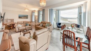 A spacious living room with leather sofas, a dining table set for four, and large windows offering a view of the sea. - 3 Bed in Woolacombe (oc-n1711) (Woolacombe)