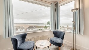 A room with two armchairs, a small round table, and a floor lamp. A large window offers a view of a beach and buildings. - 3 Bed in Woolacombe (oc-n1711) (Woolacombe)