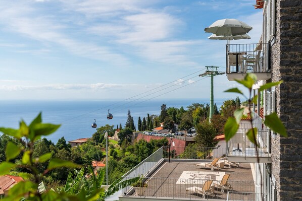 Blick auf eine Küstenlandschaft mit einer Seilbahn, einer Dachterrasse mit Liegestühlen und einem Steinhaus mit Balkon.