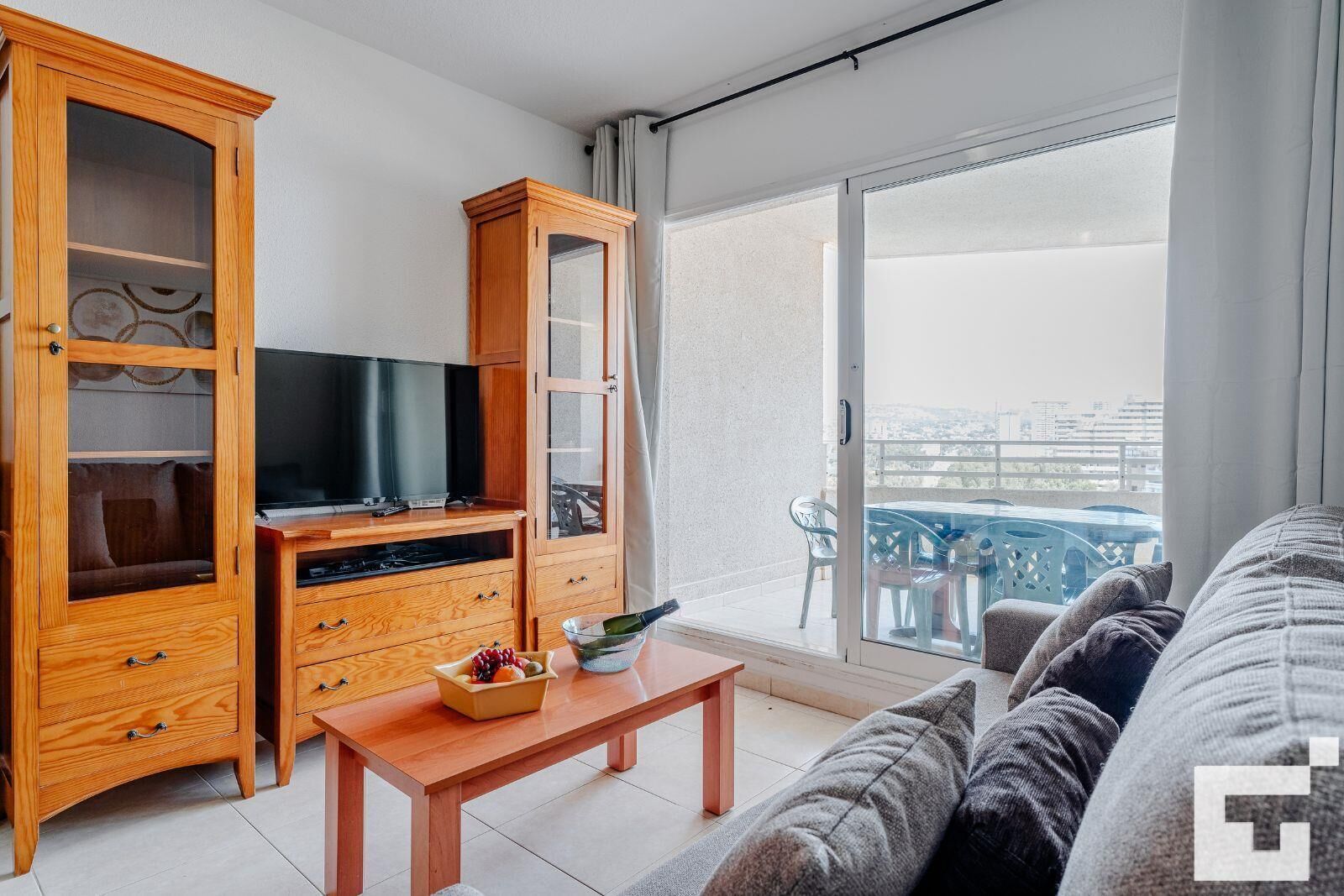 Un soggiorno moderno con un mobiletto in legno, un televisore a schermo piatto, un tavolino in vetro con una ciotola di frutta e un balcone con vista sulla città.