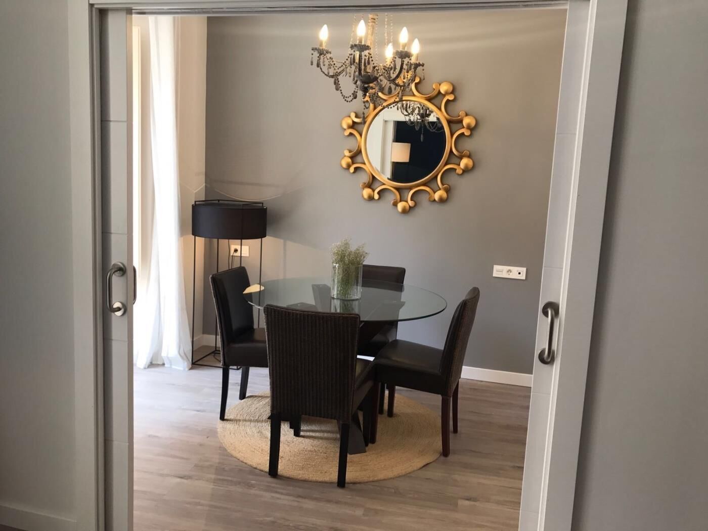 Un comedor con una mesa redonda de cristal, cuatro sillas, una araña y un espejo decorativo en la pared.