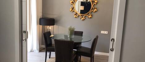 Un comedor con una mesa redonda de cristal, cuatro sillas, una araña y un espejo decorativo en la pared.
