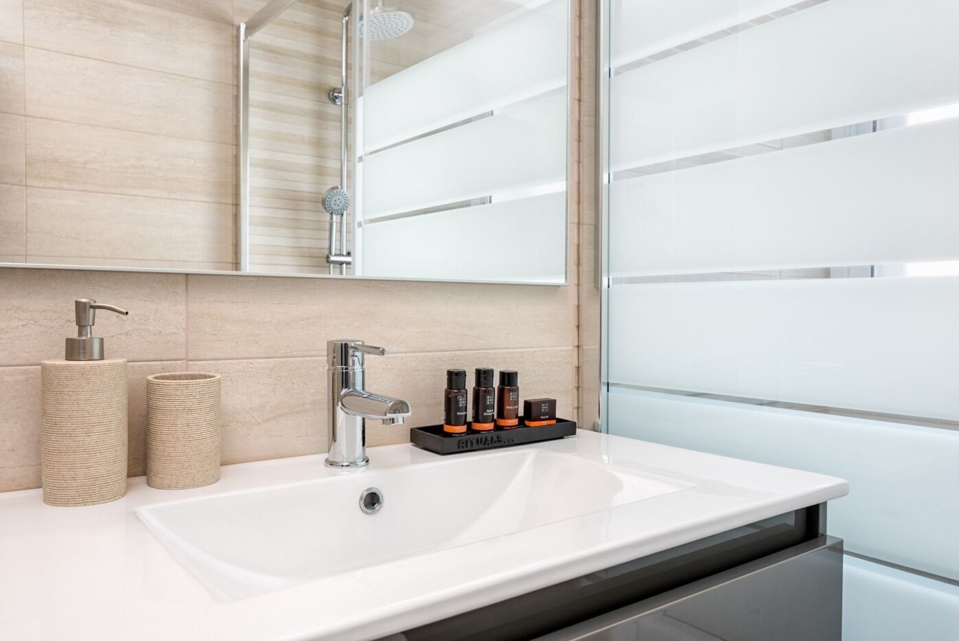 Une salle de bain moderne avec un lavabo blanc, un robinet chromé, une pomme de douche et un miroir au-dessus du lavabo. On trouve deux récipients cylindriques sur le plan de travail du lavabo.