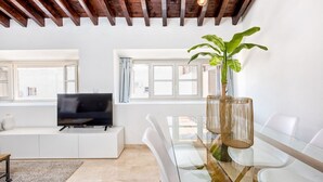 Una sala de estar moderna con un televisor sobre un mueble blanco, una mesa de comedor de vidrio con sillas y una planta grande en una maceta de fibras naturales.