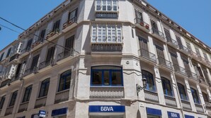 Un edificio en esquina con varios balcones, un llamativo letrero de 'BBVA' y un cielo azul despejado.