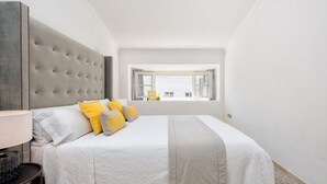 Un dormitorio con una cama grande, cabecera gris y ventana con vistas a edificios.