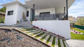 Una casa moderna de dos pisos con techo plano, un porche cubierto y un pequeño jardín con flores rojas y una terraza de madera.