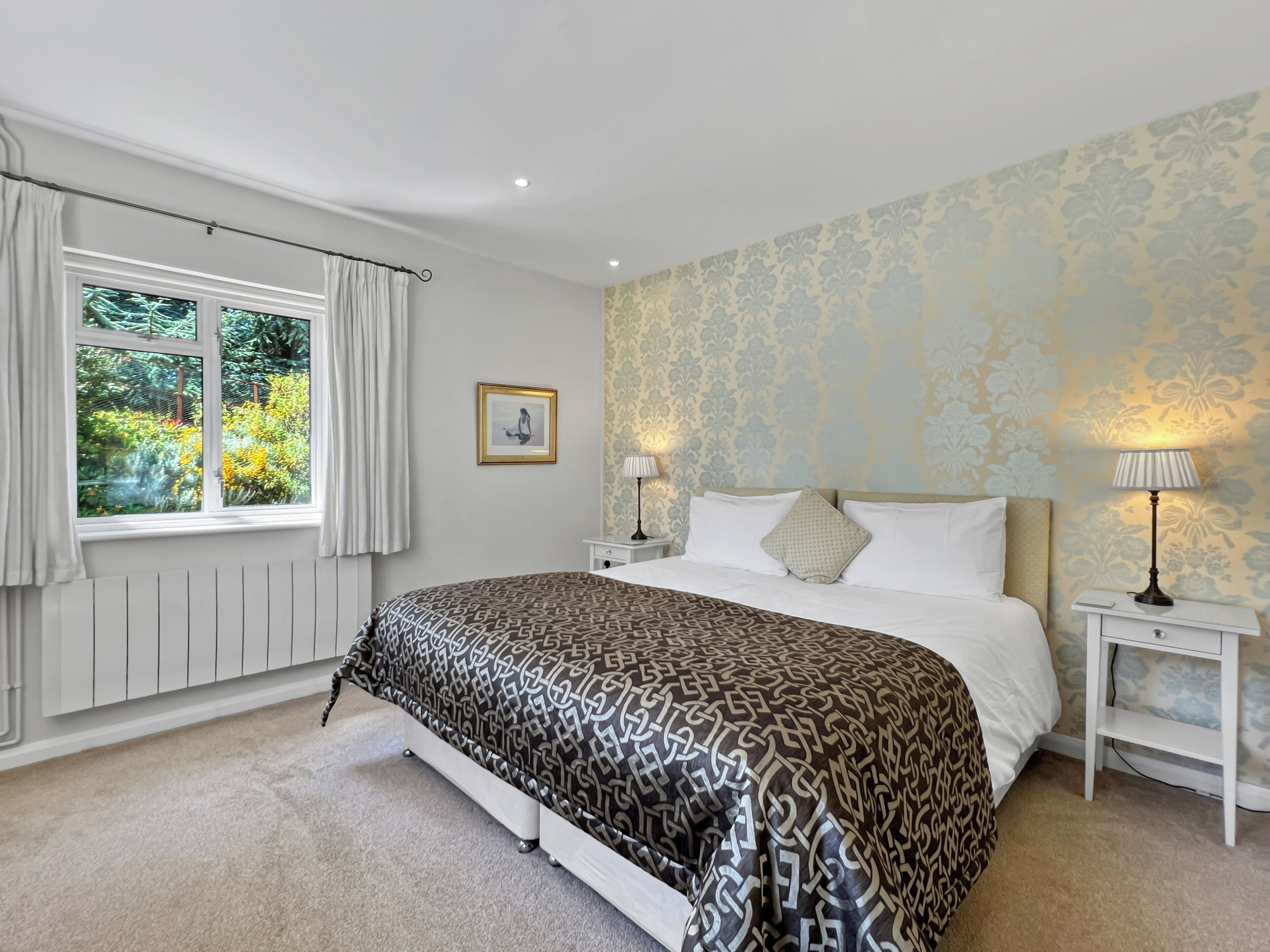 A bedroom with a large bed, two bedside tables, a window with curtains, a framed picture on the wall, and patterned wallpaper.