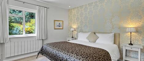 A bedroom with a large bed, two bedside tables, a window with curtains, a framed picture on the wall, and patterned wallpaper.