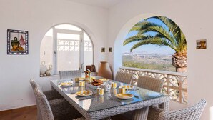 A dining area with wicker furniture, a glass table, and a scenic view through an arched window. - Villa Sabine - PlusHolidays (Benissa)