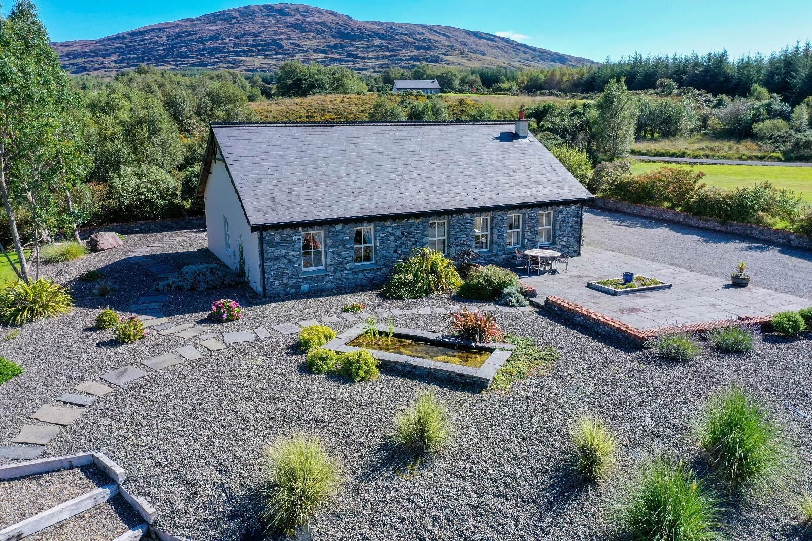 Una casa in pietra con vialetto sterrato, un piccolo giardino con uno stagno e una montagna sullo sfondo.