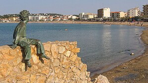 A statue of a seated figure on a stone wall by a waterfront.