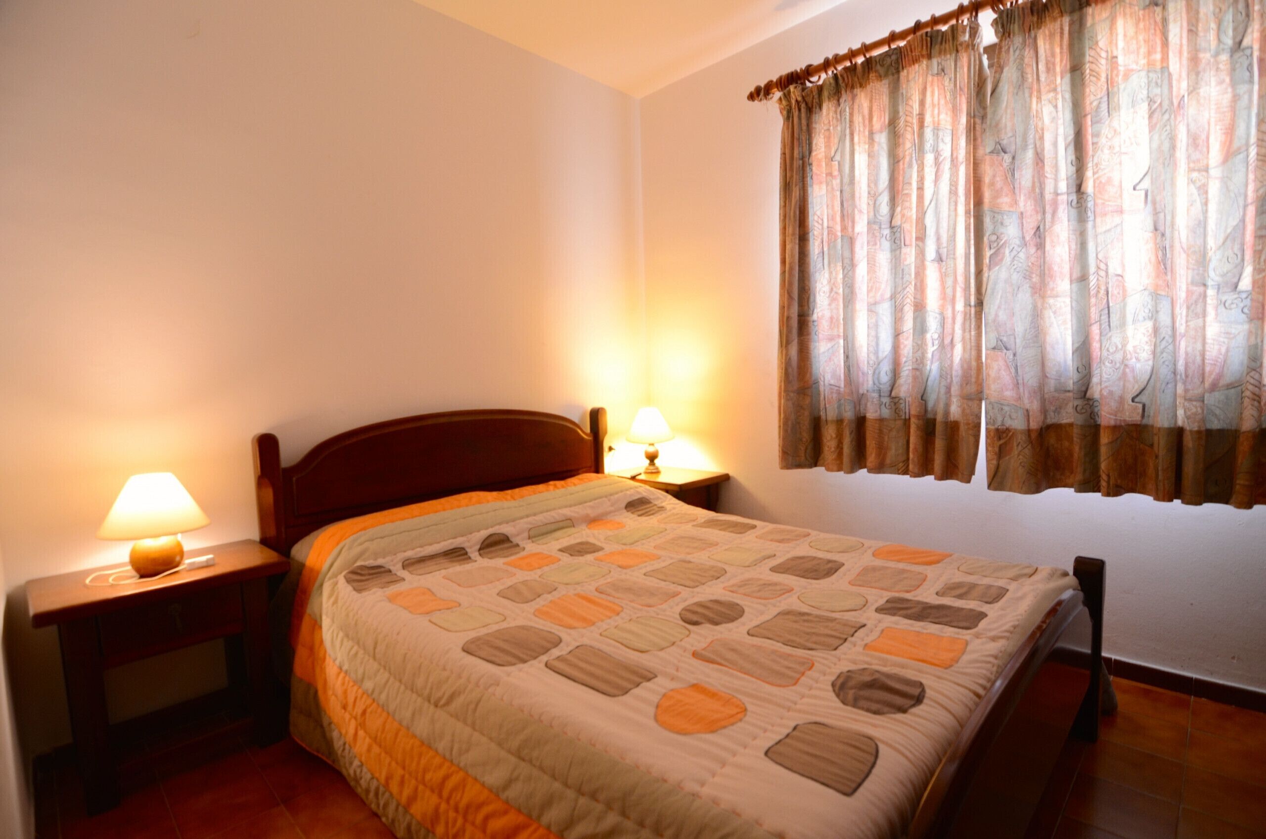 A bedroom with a bed, two bedside lamps, a window with curtains, and a wooden headboard.