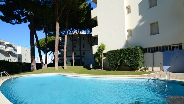 A swimming pool with a clear blue water surface, surrounded by a grassy area and trees. - APARTMENT PUEBLO AMARRES 2D (L'Escala)