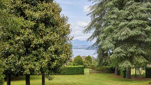 A well-maintained garden with a variety of trees, a neatly trimmed hedge, and a view of a lake with mountains in the distance. - MAJOR LAKE VILLA WITH GARDEN & SWIMMING POOL. AMAZING VIEW ON THE LAKE AND ALPS (Ranco)