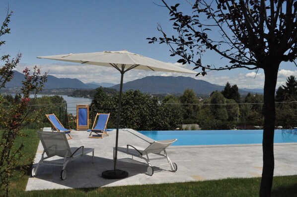 A poolside area with lounge chairs and a large umbrella, overlooking a body of water and mountains.