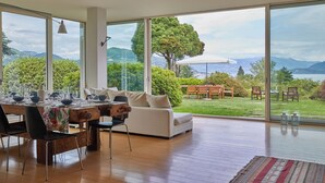 A dining area with a table set for four, a sofa, and a view of a garden with a wooden table and chairs. - MAJOR LAKE VILLA WITH GARDEN & SWIMMING POOL. AMAZING VIEW ON THE LAKE AND ALPS (Ranco)