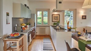 A modern kitchen with a central island, a dining area with a green tablecloth, and a view of the outdoors through large windows. - MAJOR LAKE VILLA WITH GARDEN & SWIMMING POOL. AMAZING VIEW ON THE LAKE AND ALPS (Ranco)
